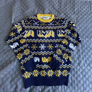 Mönstrad tröja från HV71 - Säljer en supercool tröja från HV71 med ett snyggt mönster i blått, gult och vitt. Perfekt för vintern med långa ärmar och ett mysigt stickat material. Den har ett sportigt tema med hockeyklubbor och snöflingor. Grym för att visa ditt stöd för laget!