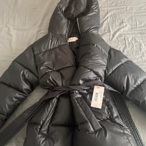 Svart pufferjacka med bälte - Säljer en snygg svart pufferjacka, som är helt ny med prislapp. Jackan har en huva och ett bälte i midjan för en stilren look. Perfekt för kalla vinterdagar. Den är långärmad och har en dragkedja framtill. Supermysig och varm!