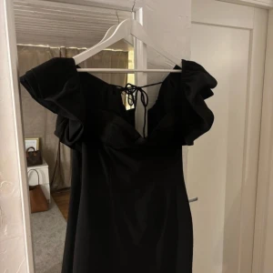 Svart klänning med volangdetaljer - Säljer en elegant svart klänning med volangdetaljer och offshoulder-design. Klänningen har en knytning baktill och är perfekt för festliga tillfällen. Storlek L