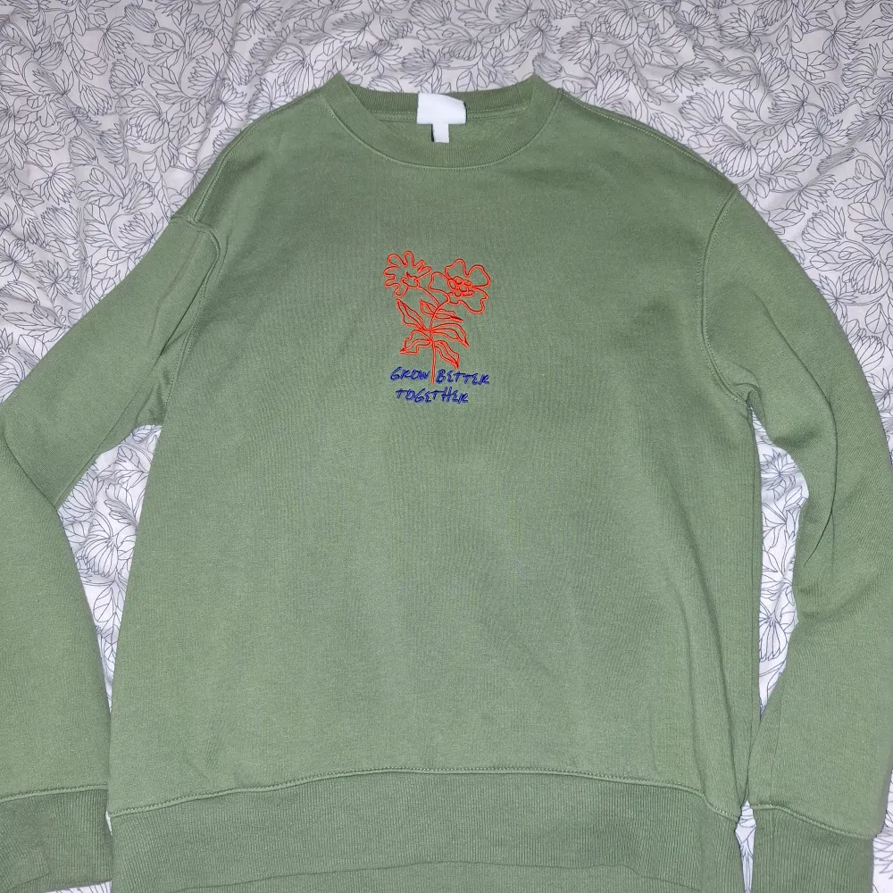 Säljer en grön sweatshirt från H&M i storlek S. Tröjan har ett coolt broderat motiv med texten 'Grow Better Together' i orange och blått på framsidan. Den är i avslappnad passform och perfekt för både vår och höst. Superbekväm och stilren!. Tröjor & Koftor.