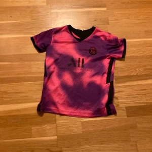 Rosa och lila fotbollströja - Säljer en cool fotbollströja i rosa och lila med korta ärmar. Tröjan har ett tryck på framsidan och Mbappé nummer 7 på baksidan. Perfekt för fotbollsfans som vill sticka ut på planen eller läktaren! 150 kr ink frakt!