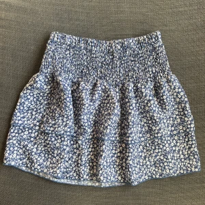 Blå blommig kjol - Superfin blå kortkjol med vitt blommönster. Den har en elastisk midja som gör den bekväm och lätt att bära. Perfekt för sommardagar eller en avslappnad vårlook. Kjolen är i mycket bra skick och passar perfekt till en enkel topp eller blus.