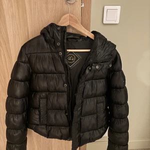 Svart pufferjacka från Superdry - Säljer en svart pufferjacka från Superdry i mycket bra skick. Jackan har en skön huva och stängs med dragkedja. Perfekt för kyliga dagar och ger en cool look. Passar bra till både vardag och fest. Storleken är 38.
