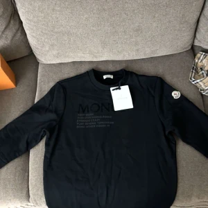 Svart tröja från Moncler - Säljer en snygg svart tröja från Moncler i mycket bra skick. Den har långa ärmar och ett stilrent tryck på framsidan. Perfekt för både vardag och fest. Moncler-loggan finns på ärmen för en extra touch av lyx. Passar perfekt till jeans eller chinos!