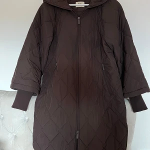 Isay Brun quiltad kappa - Säljer en supermysig brun quiltad kappa med dragkedja. Den har långa ärmar och en skön, lös passform som är perfekt för höst och vinter. Jackan har en stilren design med ribbade muddar och en liten slits på sidorna. Perfekt för att hålla sig varm och snygg under kyliga dagar!