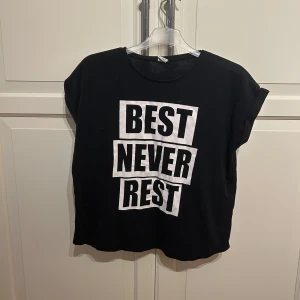 Svart t-shirt med text från Lindex - Cool svart t-shirt från Lindex med texten 'BEST NEVER REST' i stora vita bokstäver. Perfekt för träning. T-shirten har korta ärmar och är i bra skick. Passar perfekt för dig som vill ha en bekväm och stilren look.