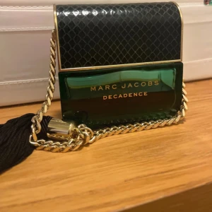 Marc Jacobs Decadence parfym - Säljer en lyxig Marc Jacobs Decadence parfym. Flaskan är grön med en elegant svart och guld design, formad som en handväska med en guldkedja och svart tofs. Perfekt för den som vill ha en doft som utstrålar glamour och stil. Flaskan är i gott skick och redo att användas.