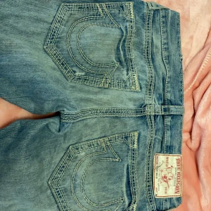 Blå jeans från True Religion - Säljer ett par snygga blå jeans från True Religion. De har en loose passform och är lågmidjade, perfekt för en avslappnad stil. Jeansen har de klassiska True Religion-sömmarna på bakfickorna och är i bra skick. Perfekta för vardagsbruk! Passar S/M. Tvättas innan utskick
