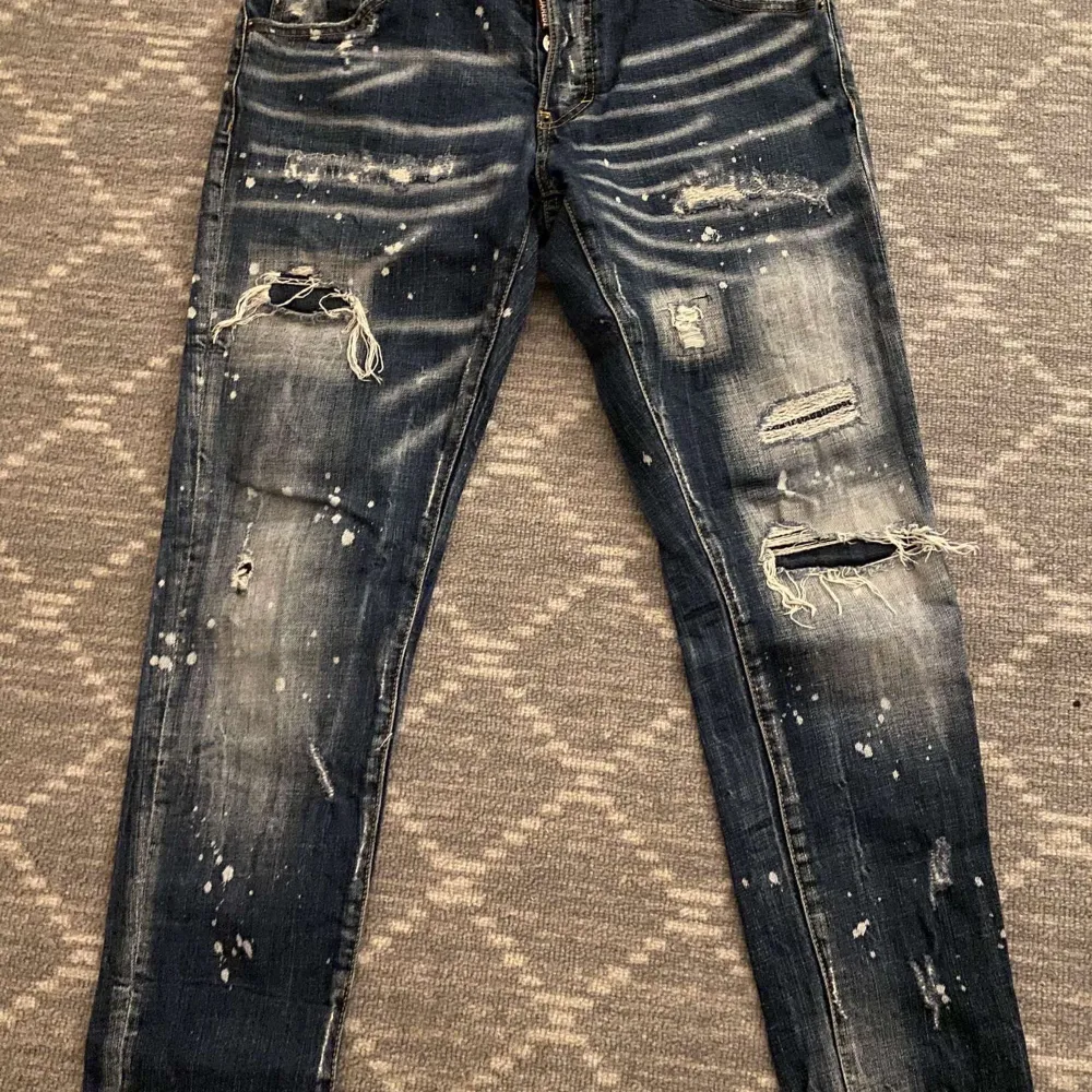 Snygga blå jeans från Dsquared2 med slitna detaljer och färgstänk. De har en klassisk femficksdesign och knappgylf. Perfekta för en trendig look. Storlek 48/S/M passar båda storlekar.. Farkut & Housut.