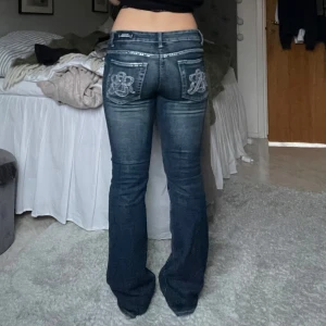Mörkblå jeans med broderade bakfickor - Snygga mörkblå jeans med broderade detaljer på bakfickorna. De har en låg midja och bootcut-stil som ger en klassisk look. Perfekta för en avslappnad stil med en touch av elegans. Supersnygga och knappt använda💕midjemåttet är 42cm, jag är 168 och dom är perfekt längd på mig, jag skulle säga att dom är längd 34. Några stenar baktill saknas, men inget man tänker på (se bild 2) 💞