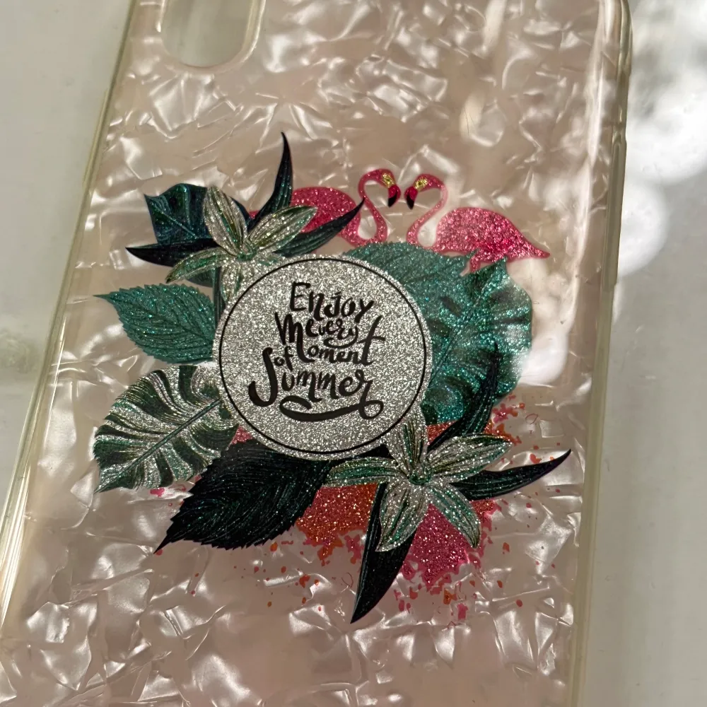 Snyggt transparent mobilskal med ett unikt mönster av flamingos och gröna blad. Texten 'Enjoy every moment summer' är tryckt i mitten med glittriga detaljer. Perfekt för att ge din mobil en somrig och stilren look. Passar iphone XR.. Asusteet.