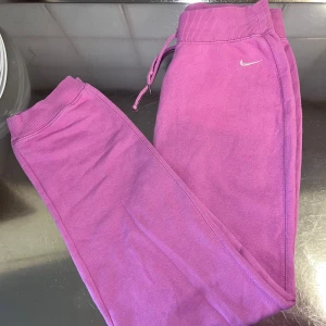 Rosa mjukisbyxor från Nike - Säljer ett par rosa mjukisbyxor från Nike med en liten vit logga på sidan. Byxorna har ribbade muddar och snörning i midjan för en bekväm passform. Perfekta för en avslappnad stil.
