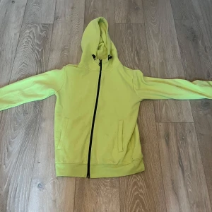 Gul hoodie med dragkedja - Säljer en fräsch gul hoodie med dragkedja och huva. Perfekt för en avslappnad stil. Den har långa ärmar och praktiska fickor framtill. Passar bra till både jeans och joggers.