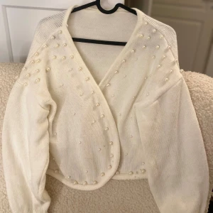 Vit kofta med pärlor - Säljer en elegant vit kofta dekorerad med pärlor. Koftan har en omlott-design och långa ärmar, vilket ger en stilren och chic look. Perfekt för att lägga till en touch av elegans till din outfit.
