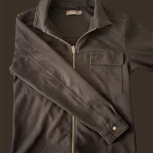 Svart jacka från Dressmann - Snygg svart jacka från Dressmann med dragkedja och krage. Jackan har långa ärmar och en bröstficka med knapp. Perfekt för en stilren look.