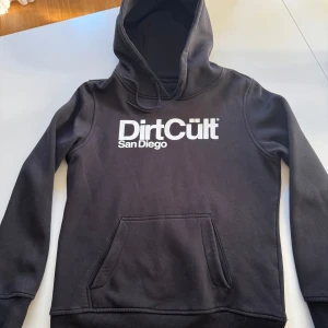 Svart hoodie från DirtCült - Säljer en svart hoodie från DirtCült med tryck 'San Diego' i vitt på bröstet. Tröjan har en klassisk känguruficka och justerbar huva med dragsko. Perfekt för en avslappnad stil.