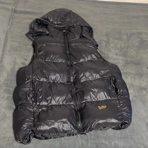 Svart dunväst med huva från Black good  - Snygg svart dunväst från Black Bank med huva och dragkedja. Västen har en glansig yta och en cool guldfärgad insida. Perfekt för kyligare dagar när du vill hålla stilen. Väldigt skön och ha på sig och värmer bra. Använd fåtal gånger men inga skador alls.