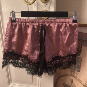 Rosa satinshorts med spetsdetaljer - Jag säljer ett par gulliga rosa shorts i satin med svarta spetsdetaljer längs nederkanten. De har en svart knytning framtill som ger en extra touch. Perfekta för en chic och bekväm look.❤️❤️