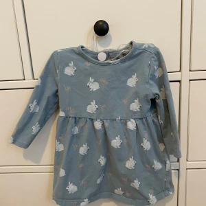 Blå klänning med kaninmönster från Newbie - Söt blå klänning från Newbie med ett gulligt kaninmönster. Klänningen har långa ärmar och en rund halsringning. Perfekt för små äventyrare som vill ha något bekvämt och stiligt på samma gång.