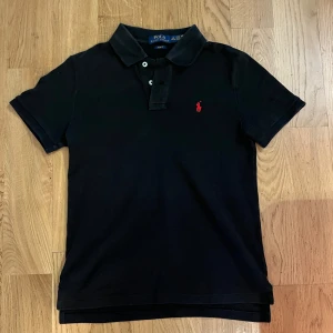 Svart pikétröja från Ralph Lauren - Säljer en stilren svart pikétröja från Ralph Lauren i slim fit. Tröjan har korta ärmar och en klassisk krage med knappar. Den är dekorerad med Ralph Laurens ikoniska röda logotyp på bröstet. Perfekt för en avslappnad men ändå elegant look.