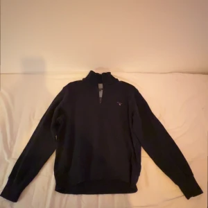 Marinblå tröja med halv dragkedja från GANT - Säljer en stilren marinblåa tröjan från GANT med halv dragkedja och broderad logotyp på bröstet. Perfekt för en avslappnad men snygg look. Tröjan har långa ärmar och en bekväm passform.