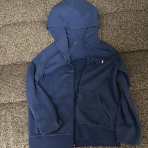 Blå Zip Hoodie från Polo Ralph Lauren - Säljer en snygg blå huvtröja från Polo Ralph Lauren. Tröjan har en dragkedja framtill och en liten broderad logga på bröstet. Perfekt för en avslappnad stil med sina långa ärmar och bekväma passform.