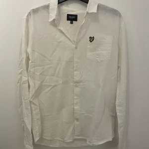 Vit skjorta från Lyle & Scott - Säljer en stilren vit skjorta från Lyle & Scott med lång ärm och klassisk krage. Skjortan har en broderad logga på bröstfickan och knappar framtill. Perfekt för en snygg och avslappnad look. Skriv om ni har några funderingar och priset är inte hugget i sten!
