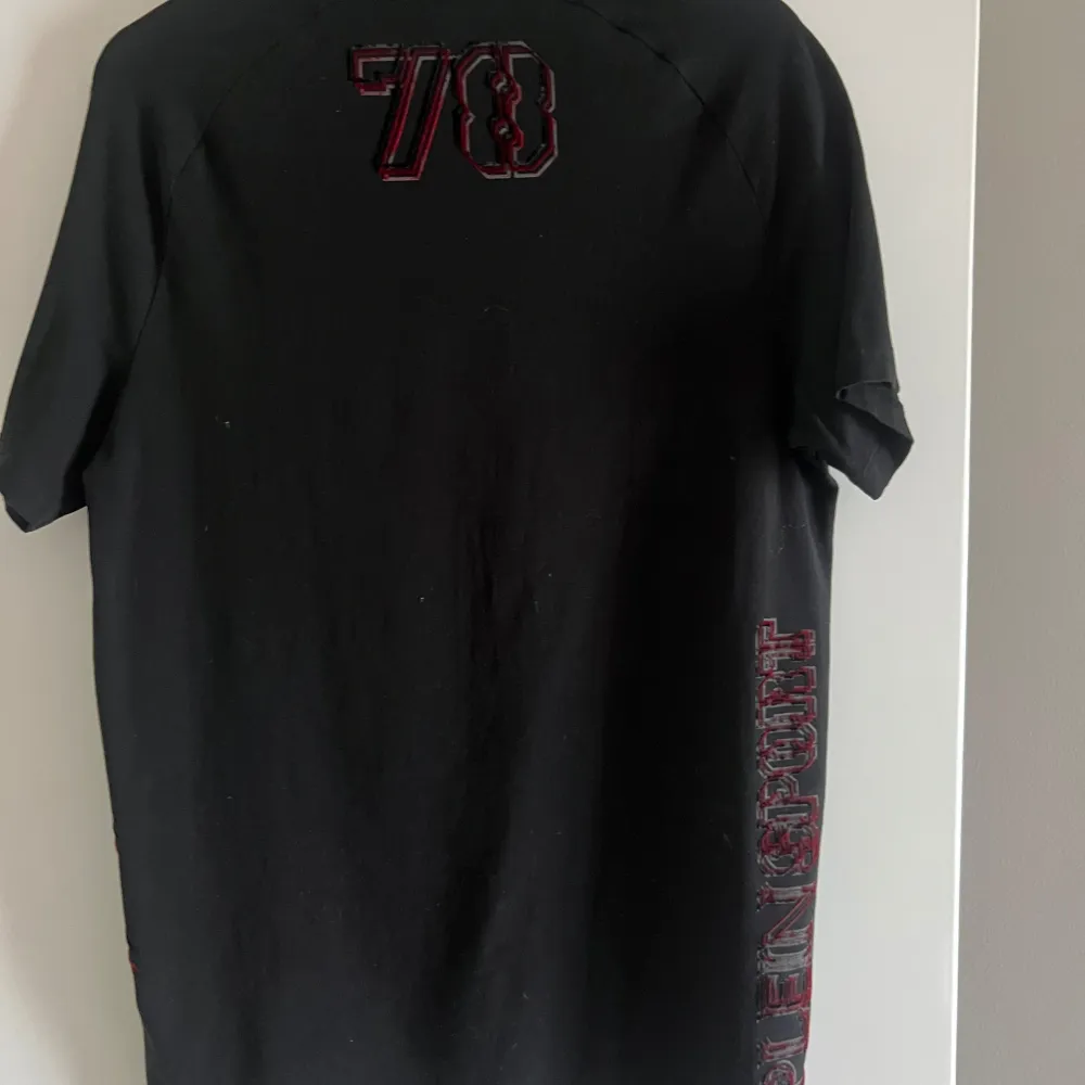 Snygg svart t-shirt från Plein Sport med coola röda detaljer och ett märke på bröstet. T-shirten har korta ärmar och ett tryck med siffran 78 på baksidan. Perfekt för en sportig look!. T-paidat.