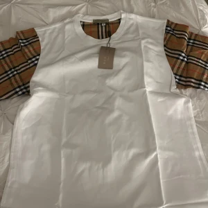 Vit t-shirt med rutiga ärmar från Burberry - Säljer en stilren vit t-shirt från Burberry med klassiska rutiga ärmar i beige, svart och röd. Perfekt för en trendig och avslappnad look. T-shirten har en normal passform och är tillverkad i mjukt bomullsmaterial. Helt ny passar l/m
