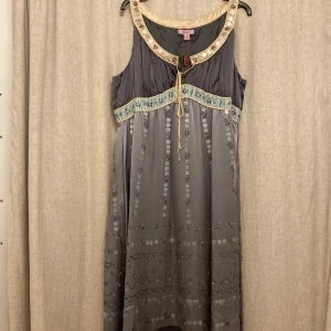 Grå klänning från Monsoon - Elegant grå klänning från Monsoon med vackra broderade detaljer och dekorativa paljetter. Klänningen har en knytning framtill och en mjuk, flödande design. Perfekt för en stilfull look.