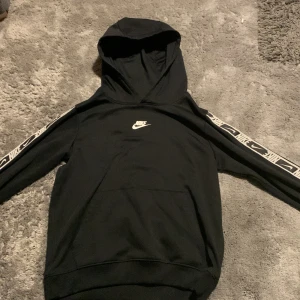 Svart hoodie från Nike - Säljer en svart Nike hoodie med Dri-FIT teknologi. Den har en klassisk design med Nike-logga på bröstet och mönstrade band längs ärmarna. Perfekt för en sportig look eller avslappnad stil. 🖤