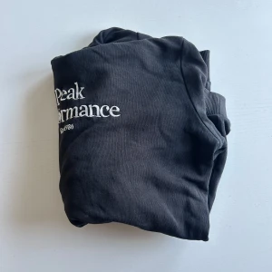 Svart hoodie från Peak Performance - Säljer en svart hoodie från Peak Performance med vit logga på bröstet. Tröjan har en klassisk känguruficka. Perfekt för en avslappnad stil. Hör av er vid minsta fundering!