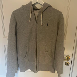 Grå hoodie från Polo Ralph Lauren - Säljer en stilren grå hoodie från Polo Ralph Lauren med dragkedja och justerbar huva. Tröjan är i mycket gott skick och den har använts varsamt.