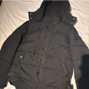 Jacka - Säljer en svart pufferjacka från SMOG med huva och dragkedja. Jackan har knappar och flera praktiska fickor. Perfekt för kyliga dagar.