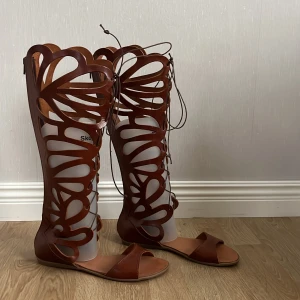 Bruna sandaler med snörning - Hantverk. Snygga bruna sandaler med högt skaft och unikt utskuret mönster. De har snörning framtill och dragkedja bak för enkel påtagning. Köpta för 800kr och knappt använda. 