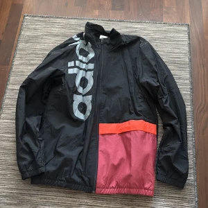 Svart vindjacka från Adidas - Snygg svart vindjacka från Adidas med stor logga på framsidan. Jackan har en dragkedja och en cool färgblockdesign med inslag av rött och orange. Perfekt för en sportig look.