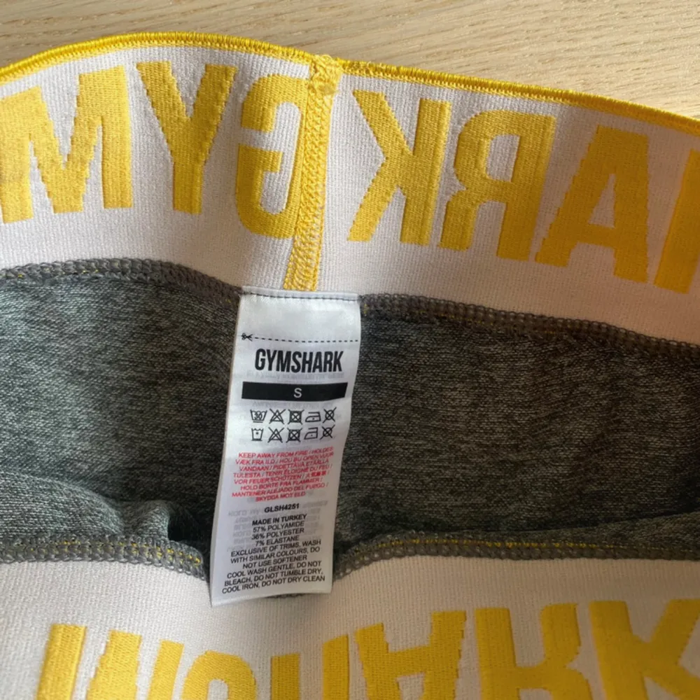 Snygga grå träningsshorts från Gymshark med en elastisk gul midja där märkesnamnet är tryckt i vitt. Perfekta för gymmet eller löprundan. De är designade för att sitta bekvämt och ge bra rörelsefrihet.. Sportswear.