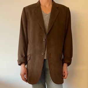 Brun kavaj från Madison Avenue - Säljer en stilren brun mockaliknande kavaj från Madison Avenue. Kavajen har en klassisk design med två knappar framtill och långa ärmar. Perfekt för en elegant look.