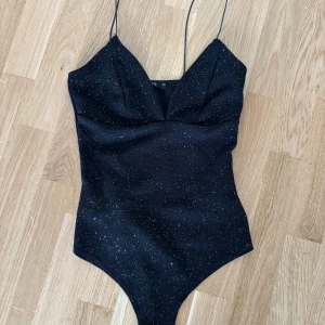 Svart glittrig bodysuit från NLY One - Snygg svart bodysuit från NLY One med glittriga detaljer. Den har tunna axelband och en figurnära passform som ger en elegant look. Perfekt för en kväll ute eller en speciell tillställning.