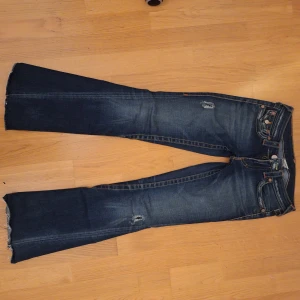 Mörkblå True Religion - Snygga mörkblå jeans från True Religion med slitna detaljer och bootcut-stil. De har dekorativa bakfickor med knappar och en klassisk femficksdesign. Perfekta för en avslappnad look. Skriv vid frågor och prisförslag💕 Midja: 38 innerbenslängd: 77 ytterbenslängd: 95. 