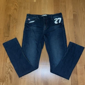 Mörkblå jeans från Hampton Republik  - Snygga mörkblå jeans med ett '27' tryck på fickan. De har en klassisk femficksdesign och en knappgylf. Perfekta för en avslappnad stil med en touch av unik detalj. Storlek 170cm.