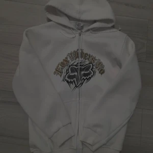Vit hoodie från Fox Racing  - Säljer en vit hoodie med tryck från Monster Energy och Fox Racing. Den har en dragkedja framtill och en stor logga på ryggen. 
