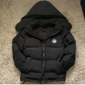 Svart pufferjacka från Trapstar - Säljer en svart pufferjacka från Trapstar i mycket bra skick. Jackan har en dragkedja framtill och en avtagbar huva. Den är perfekt för kalla dagar och har en cool logga på bröstet och text på ryggen. Passar perfekt för höst och vinter!