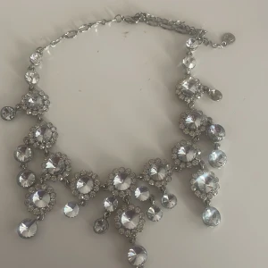 Glittrigt halsband med stora stenar - Säljer ett fantastiskt glittrigt halsband i silverfärg med stora, klara stenar som fångar ljuset perfekt. En sten fattas.Perfekt för fest eller speciella tillfällen. Halsbandet har en justerbar kedja för att passa olika storlekar. Ett riktigt statement-smycke som drar blickarna till sig!