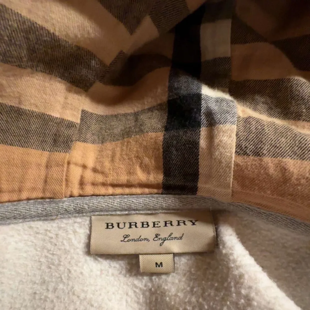 Säljer en stilren grå hoodie från Burberry i storlek M. Den har en dragkedja framtill och en klassisk rutig insida i huvan. Perfekt för både vardag och lite kyligare dagar. Passar bra till jeans eller joggers. Hör av dig vid intresse!. Tröjor & Koftor.