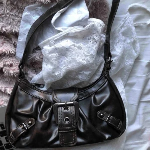 Svart axelväska med spänne - Snygg svart axelväska i skinn med coola spänndetaljer framtill. Perfekt för både vardag och fest! Den har en justerbar axelrem och är i mycket bra skick. En stilren och praktisk väska som passar till alla outfits. Köpt för 900, säljer för 500