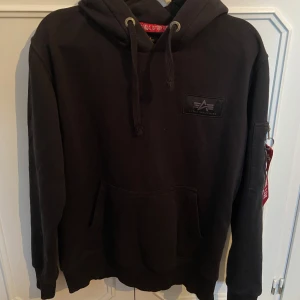 Svart hoodie från Alpha Industries - Säljer en svart hoodie från Alpha Industries i mycket bra skick. Den har en stor logga på ryggen och en cool 'Remove Before Flight'-detalj på ärmen. Perfekt för höst och vinter, med en skön passform och känguruficka framtill. Huvan har dragsko för justering. Perfekt för en casual look! 