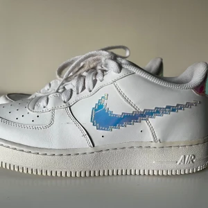 Nike Air Force med pixelmönster - Säljer ett par vita Nike Air Force med unikt pixelmönster på swooshen. Skorna är i bra skick och har en cool holografisk effekt som skiftar i blått och lila. Perfekta för att sticka ut i mängden! De har klassisk snörning och är superbekväma för vardagsbruk.