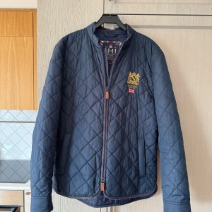 Mörkblå quiltad jacka från Morris - storlek L - Säljer en snygg mörkblå quiltad jacka från Morris i storlek L. Jackan har en broderad logga på bröstet och en dragkedja framtill. Perfekt för höst och vår med sin stilrena design och bekväma passform. Passar både till vardags och lite finare tillfällen. 🌟