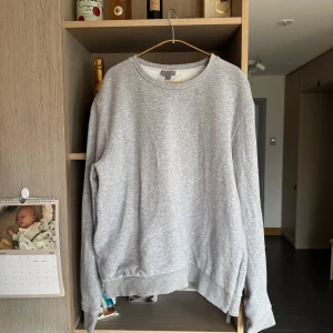 Grå Sweatshirt COS - Storlek L - Säljer en grå sweatshirt från COS i storlek L. Tröjan har en unik dragkedjedetalj på nedre sidan och är i fint skick. Perfekt för en minimalistisk och modern stil.